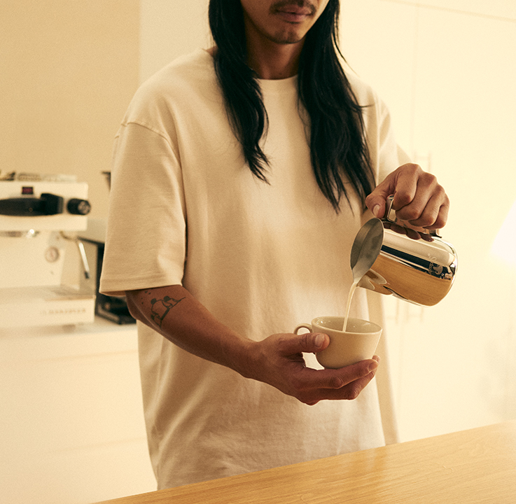 A person with tattooed arms pouring milk into a coffee cup on a home kitchen bench, warm cream tones